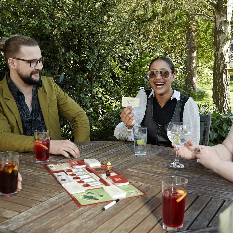 Two friends playing the Taskmaster game outside  holding a Garden Task card in the sun