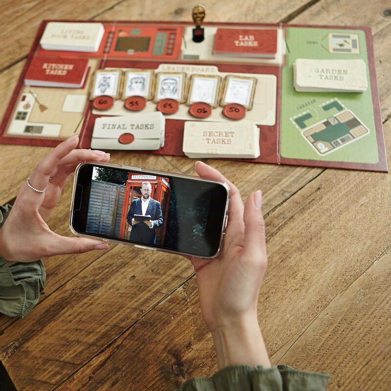 The Taskmaster Board game board and contents in the background with hands holding a phone screen showing Little Alex Horne